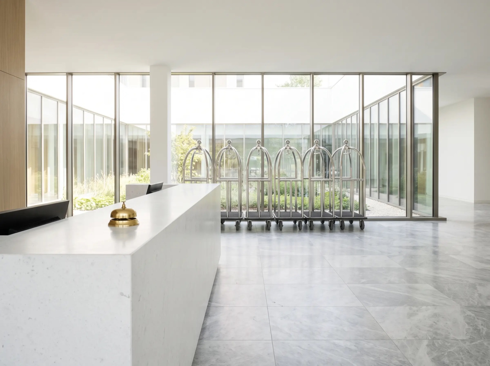 An empty hotel lobby: a long white reception counter with a brass bell and a row of luggage carts by the windows